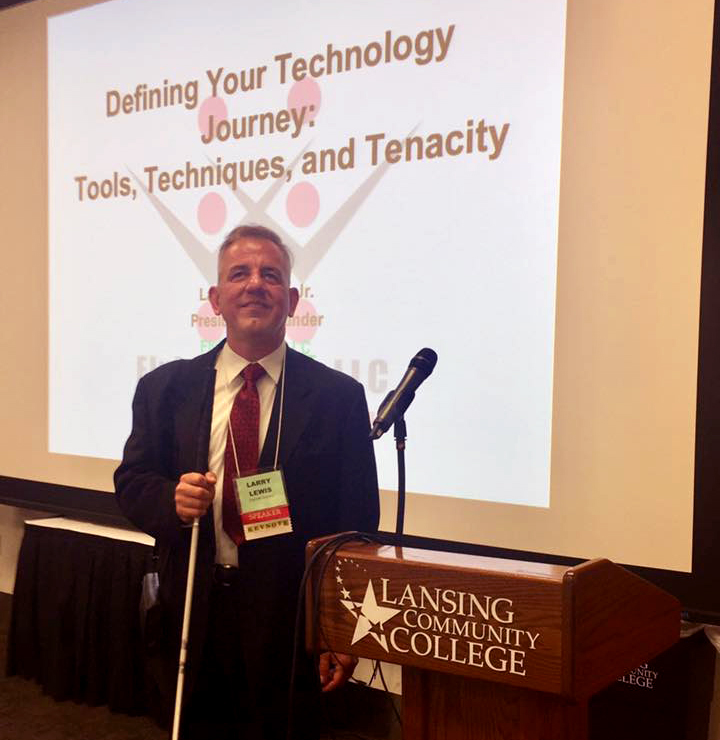 Flying Blind, LLC's Founder, Larry Lewis, in suit and tie, stands smiling in front of a large, wall-sized projection of the Flying Blind, LLC Logo, upon which are the words, 'Defining Your Technology Journey: Tools, Techniques, and Tenacity'. To Larry's left is a podium emblazoned with the logo for Lansing Community College.